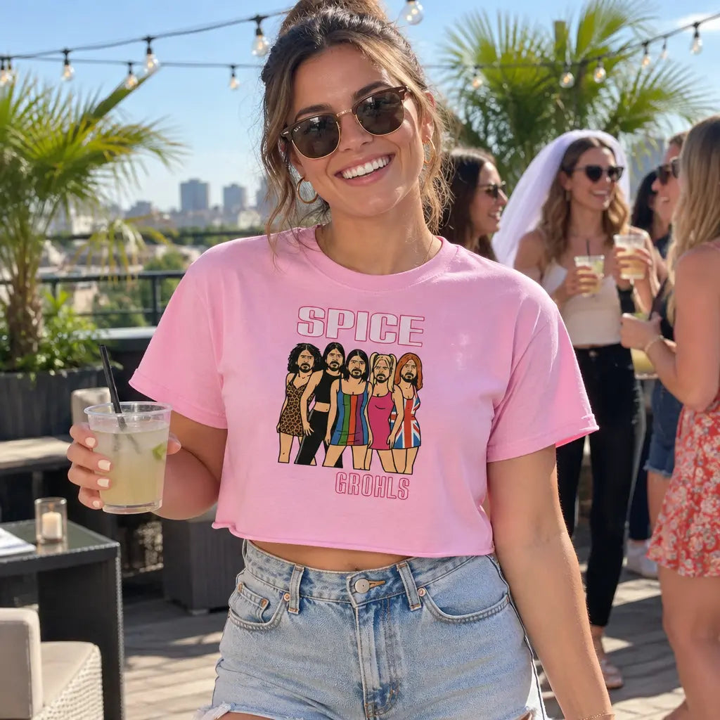 Woman at a rooftop bachelorette party holding a drink, wearing a pink Spice Grohls crop top, with the bride in a veil behind her