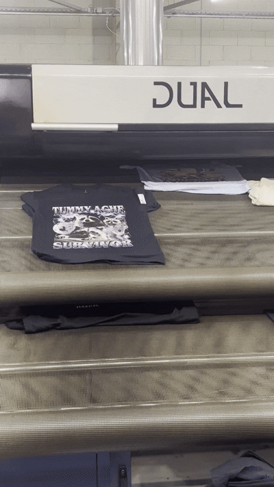Freshly printed Tummy Ache Survivor T-Shirt with raccoon graphic on the production line conveyor belt.