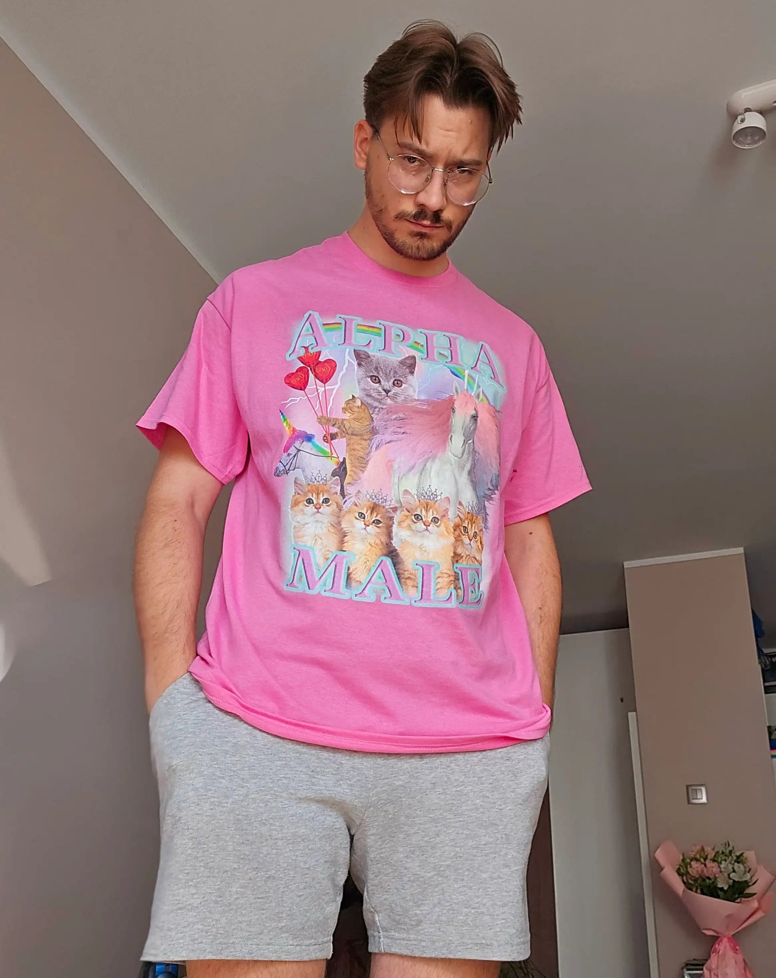 Man wearing a pink graphic tee with the text ALPHA MALE featuring an ironic design of cute kittens and a unicorn.